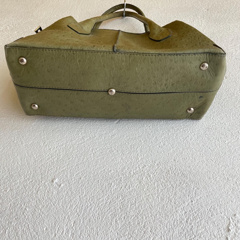 May include: Olive green handbag with a textured surface, silver-tone feet, and a top handle. The bag has a structured shape and appears to be made of leather. The bag is suspended against a white wall.