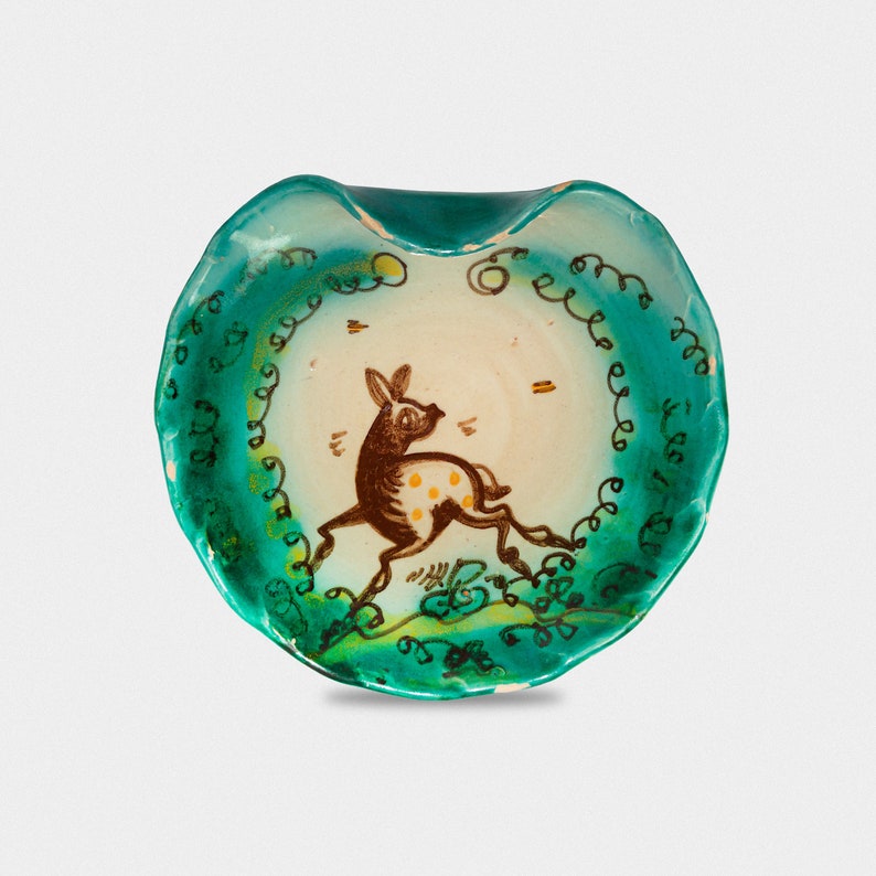 Puede incluir: Un peque&ntilde;o plato de cer&aacute;mica en forma de coraz&oacute;n con un esmalte verde. El plato presenta un dise&ntilde;o pintado a mano de un ciervo marr&oacute;n con manchas blancas, rodeado de hojas verdes y espirales.