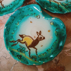 Puede incluir: Un plato de cer&aacute;mica azul turquesa con un centro blanco y un dise&ntilde;o pintado a mano de un ciervo marr&oacute;n y amarillo. El plato tiene un borde festoneado y un patr&oacute;n de enredaderas verdes alrededor del borde.