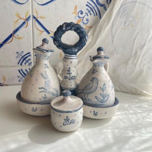 Könnte beinhalten: Ein Set aus vier Keramik-Küchenartikeln in Hellbeige mit blauen Akzenten. Das Set umfasst zwei Flaschen, ein kleines Glas und einen Griff. Die Flaschen und das Glas zeigen handgemalte Illustrationen eines Hasen und eines Vogels.