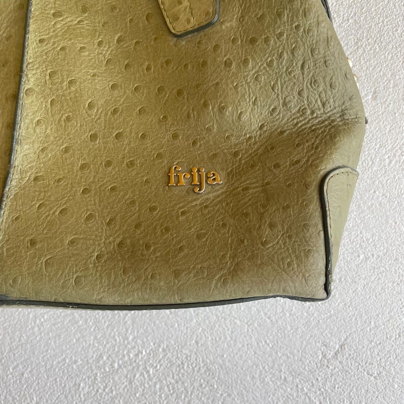 May include: Close-up of a light olive green handbag with a textured surface. The bag features the gold-coloured word "frija" on the front. The bag has a handle and a grey trim.