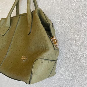May include: An olive green handbag with a textured finish, featuring a top handle and a gold-coloured clasp. The bag has a structured design with a zip closure and a small gold logo. The bag is hanging against a white wall.