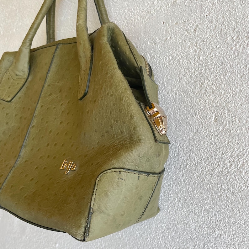 May include: An olive green handbag with a textured finish, featuring a top handle and a gold-coloured clasp. The bag has a structured design with a zip closure and a small gold logo. The bag is hanging against a white wall.