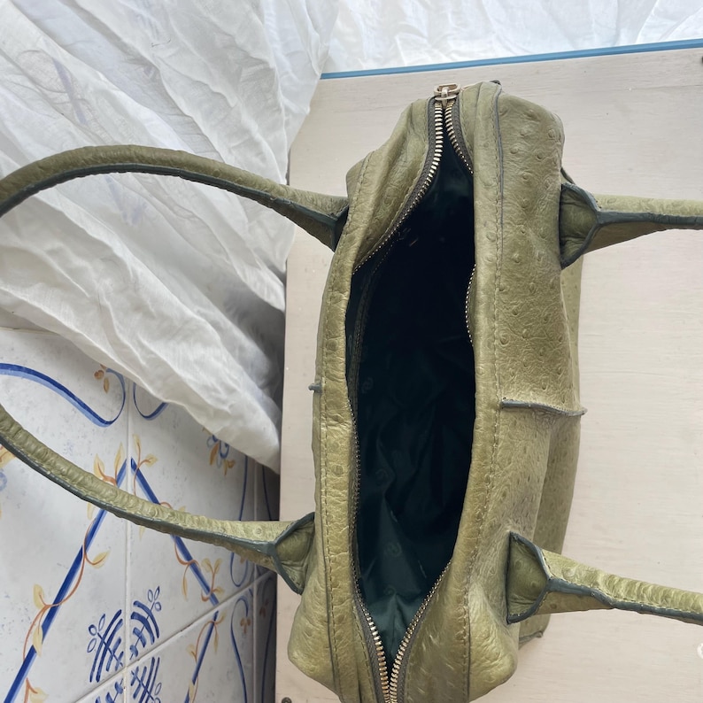 May include: Olive green leather handbag with a textured finish. The bag features a zip closure and two handles. The interior is lined with dark green fabric.