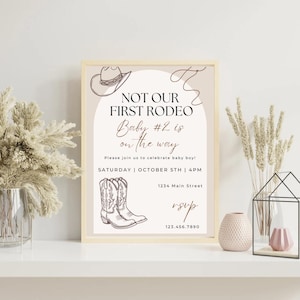 May include: A framed invitation with a beige background and a western theme. The text reads "NOT OUR FIRST RODEO Baby #2 is on the way." Includes illustrations of a cowboy hat and boots. The invitation is for a baby boy celebration.