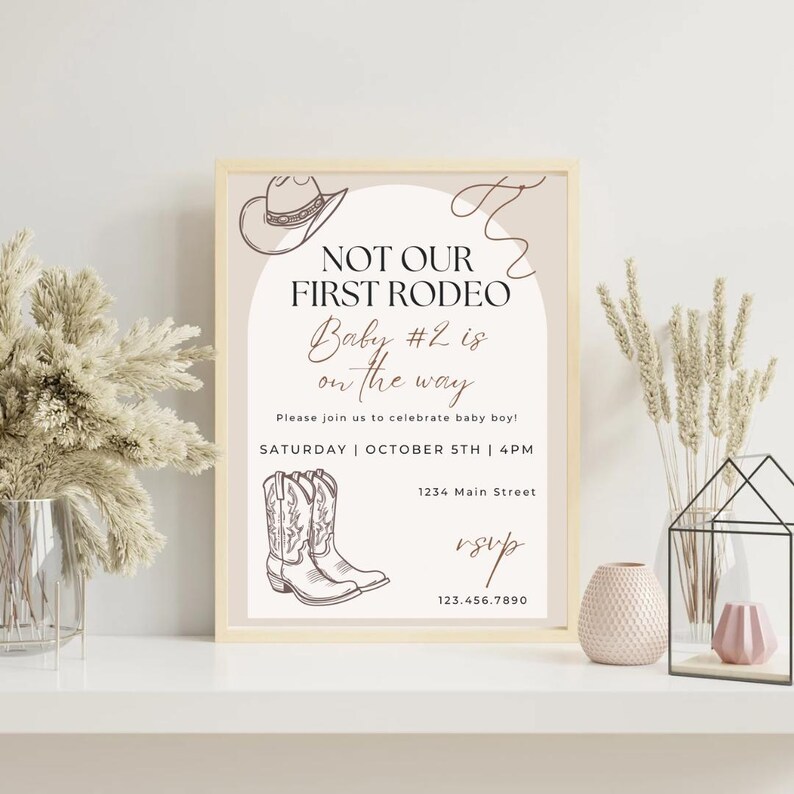 May include: A framed invitation with a beige background and a western theme. The text reads "NOT OUR FIRST RODEO Baby #2 is on the way." Includes illustrations of a cowboy hat and boots. The invitation is for a baby boy celebration.