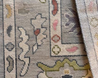 May include: Close-up of a grey rug with a floral pattern. The rug features a border with repeating floral and geometric designs in pink, green, and cream colours. The rug's texture is visible, showing the woven construction.