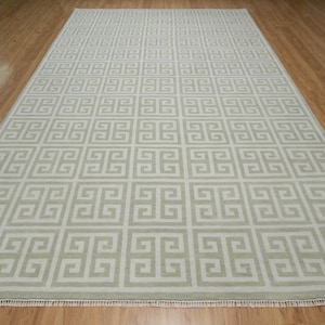 May include: A beige area rug with a white Greek key pattern. The rug has a fringe border.