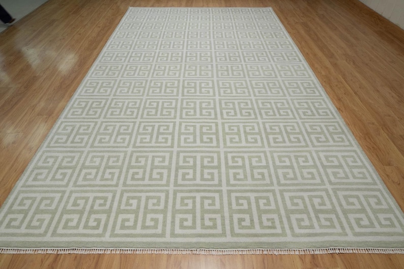 May include: A beige area rug with a white Greek key pattern. The rug has a fringe border.