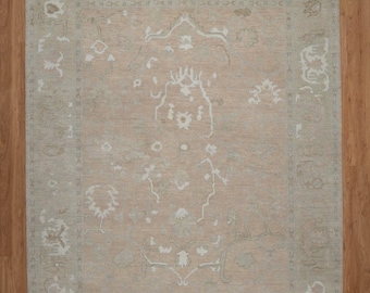 May include: A rectangular area rug with a central peach-colored field and a light green border. The rug features a detailed, ornate design in white and light green, with floral and geometric patterns. The rug is placed on a wooden floor.