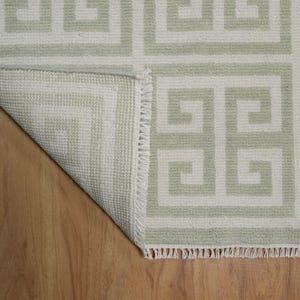 May include: A white and light green rug with a geometric pattern. The rug has a fringe border and is folded over on a wooden floor.