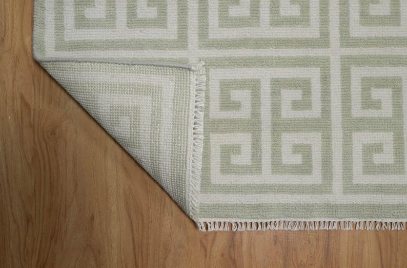 May include: A white and light green rug with a geometric pattern. The rug has a fringe border and is folded over on a wooden floor.