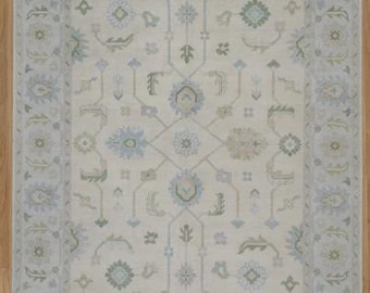 May include: Cream-coloured rectangular area rug with a border in shades of light blue and green. The rug features an intricate floral and geometric pattern, creating a traditional aesthetic. The rug is placed on a light wood floor.