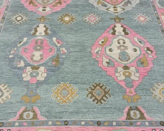 May include: A rectangular rug with a pale blue field and a repeating pattern of pink and grey geometric shapes. The rug features a pink border and gold and white accents.