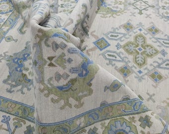 May include: A rolled-up, cream-coloured rug with a detailed pattern in shades of blue, green, and beige. The rug features geometric and floral motifs, with a border design. The texture of the rug is visible, suggesting a woven construction.