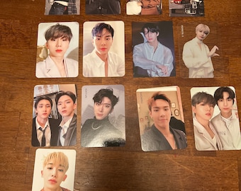 Puede incluir: Una colección de tarjetas fotográficas con un grupo de chicos de K-pop. Las tarjetas están impresas con imágenes de los miembros del grupo en varias poses y atuendos. Las tarjetas están dispuestas en una cuadrícula sobre una superficie de madera.