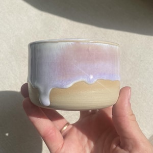 Könnte beinhalten: Ein kleiner Keramikbecher in zylindrischer Form mit einer glatten, unglasierten beigen Basis und einer glänzenden, irisierenden lavendel-rosa Glasur, die vom Rand herabläuft und einen deutlichen Schichteffekt auf der Außenfläche erzeugt.