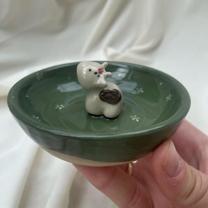 May include: A small, hand-painted ceramic dish with a dark green interior and a cream-colored exterior. The dish features a small, white cow figurine with a brown spot and a red flower. Tiny white flowers are painted around the interior.