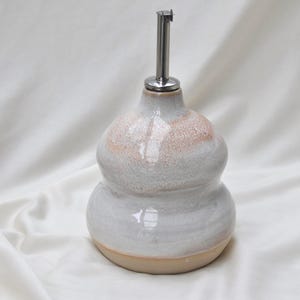 Könnte beinhalten: Ein Keramik-Ölkännchen mit bauchiger, zweistufiger Form und glänzender weißer Glasur mit subtilen pfirsichfarbenen Sprenkeln im oberen Bereich. Das Gefäß ist mit einem Ausgießer aus Edelstahl versehen, und der Boden zeigt einen unglasierten Rand.