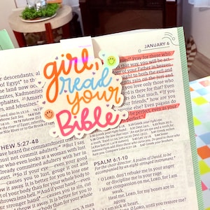 May include: A colorful die-cut sticker with the words "girl read your Bible" in orange, blue, and pink script, with smiley faces. The sticker is placed on an open Bible with highlighted text.