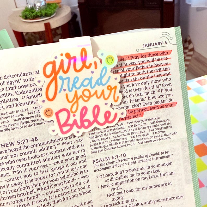 May include: A colorful die-cut sticker with the words "girl read your Bible" in orange, blue, and pink script, with smiley faces. The sticker is placed on an open Bible with highlighted text.