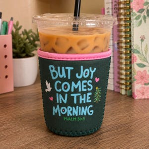 May include: A dark green neoprene iced coffee cup sleeve with a pink top rim. The text reads "BUT JOY COMES IN THE MORNING" in light blue, with a white dove, small pink hearts, and a small green Christmas tree graphic. It includes "PSALM 30:5".