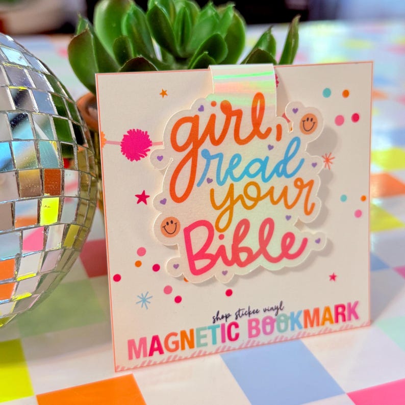 May include: A magnetic bookmark with the text "girl read your Bible" in orange, blue, and pink. The bookmark has a white background with colorful confetti and stars. The words "MAGNETIC BOOKMARK" are at the bottom.