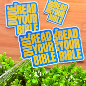 May include: Several stickers with the text "READ YOUR BIBLE DUDE" in yellow block letters on a blue background. The stickers are scattered on a wooden surface with green foliage in the foreground.