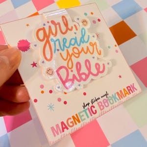 May include: A magnetic bookmark with the text "girl read your Bible" in orange, blue, and pink. The bookmark has a clear plastic overlay and is decorated with small stars and smiley faces. The bottom of the bookmark reads "MAGNETIC BOOKMARK".