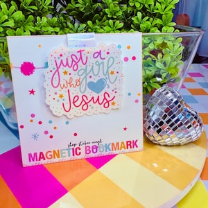 May include: A white magnetic bookmark with the text "just a girl who loves Jesus" in colorful script. The bookmark has a scalloped edge and a blue heart. The words "shop stickee vinyl" and "MAGNETIC BOOKMARK" are also printed on the bookmark.