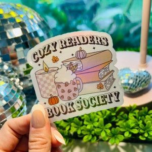 May include: A clear sticker with a fall-themed design. It features a stack of books, a candle, a cup of hot cocoa, and pumpkins. The text reads "Cozy Readers Book Society".