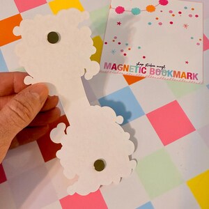 May include: White magnetic bookmark with a cloud-like design, held against a colorful checkered background. The bookmark has a small, round magnet. A square card with the text "MAGNETIC BOOKMARK" is also visible.
