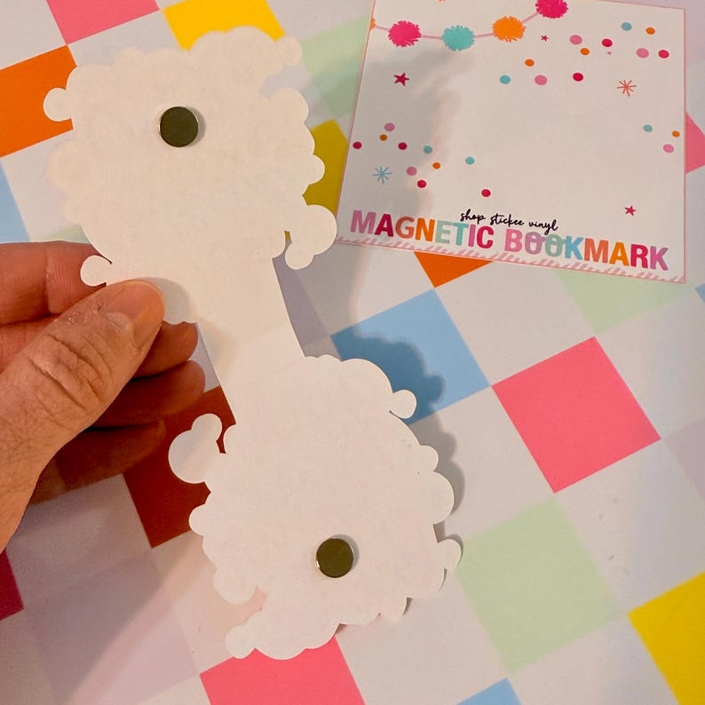 May include: White magnetic bookmark with a cloud-like design, held against a colorful checkered background. The bookmark has a small, round magnet. A square card with the text "MAGNETIC BOOKMARK" is also visible.