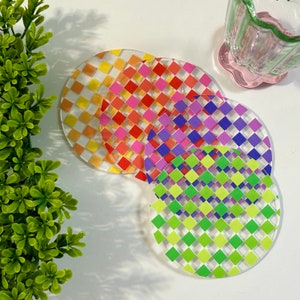 May include: Four round coasters with a checkered pattern. The coasters are layered on top of each other, with the top coaster being green and yellow, the next being purple and white, the next being red and white, and the bottom coaster being yellow and white.