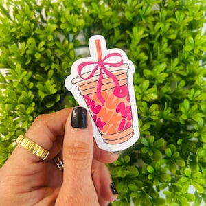 May include: A sticker of a pink and orange iced coffee cup with a pink bow and a straw. The cup has a white background and is surrounded by green foliage.