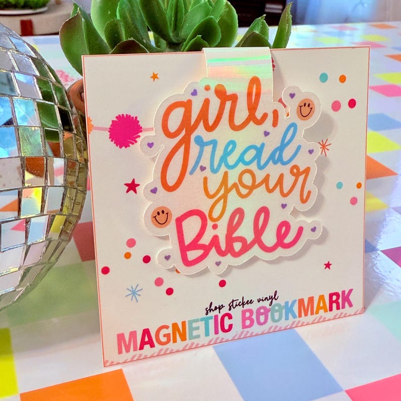 May include: A white magnetic bookmark with the text "girl read your Bible" in colorful script. The bookmark has a holographic strip and is decorated with stars, hearts, and dots. The bottom of the bookmark reads "MAGNETIC BOOKMARK".