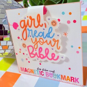 May include: A white magnetic bookmark with the text "girl read your Bible" in orange, blue, and pink. The bookmark has a decorative design with small stars and dots. The words "MAGNETIC BOOKMARK" are at the bottom.