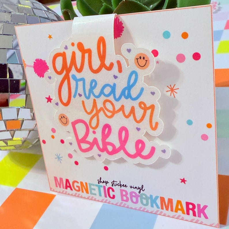 May include: A white magnetic bookmark with the text "girl read your Bible" in orange, blue, and pink. The bookmark has a decorative design with small stars and dots. The words "MAGNETIC BOOKMARK" are at the bottom.