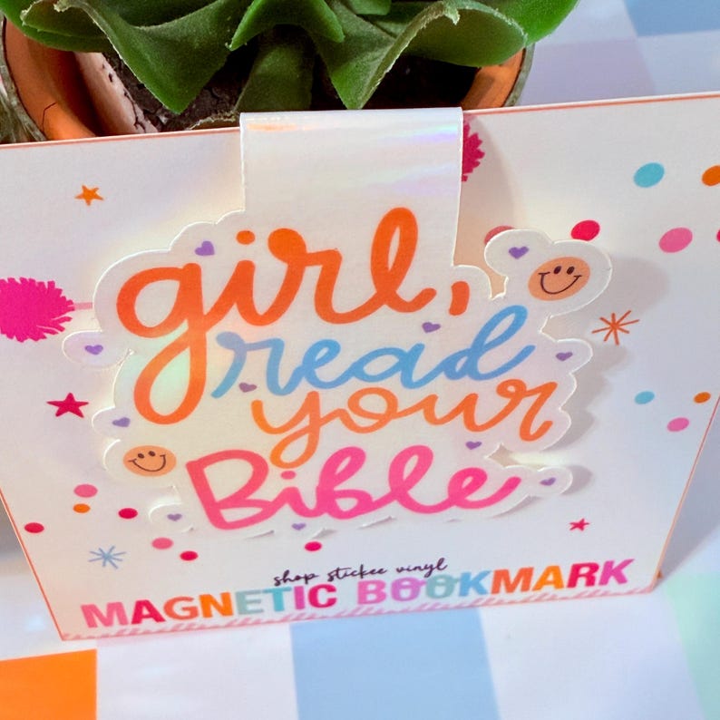May include: A white magnetic bookmark with the words "girl, read your Bible" in orange, blue, and pink. The bookmark has a holographic top and is decorated with stars and dots. The text "MAGNETIC BOOKMARK" is at the bottom.