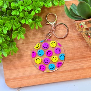 May include: A clear acrylic keychain with a gold-colored clasp and ring. The keychain features a circular design filled with colorful smiley faces in yellow, pink, and blue. The background is a wooden surface.