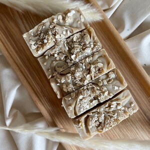 May include: A stack of six handmade oatmeal soap bars on a wooden surface. The soap bars are a light brown color and have a speckled appearance from the oatmeal.