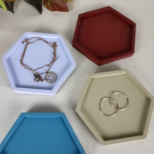 May include: Three hexagonal ceramic trinket dishes in different colours: light blue, beige, and burgundy. The dishes are on a white fabric background.