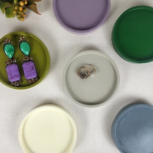 May include: Five round, decorative concrete trays in various colors. The trays are in shades of green, purple, gray, beige, and cream. The trays are perfect for holding jewelry, keys, or other small items.