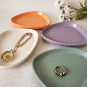 May include: Four small, irregularly shaped ceramic dishes in orange, white, purple, and green. A gold necklace with a pendant and a ring are displayed on two of the dishes. The dishes are arranged on a white surface, with white flowers in the background.