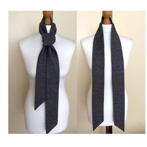 May include: A dark gray scarf is displayed on a white mannequin. The scarf is long and narrow, and it is tied in a simple knot at the neck.