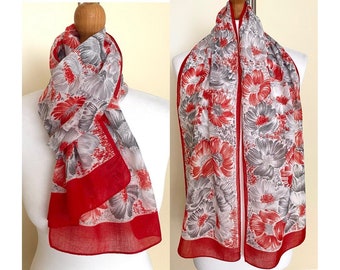 May include: A patterned scarf featuring a floral design in red and grey, with a vibrant red border. The scarf is displayed on a mannequin, highlighting its drape and fabric. The scarf is made of a lightweight material.