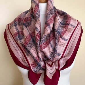 May include: A square scarf with a plaid pattern in shades of pink, red, and navy blue. The scarf has a solid burgundy border and is draped over a mannequin head. The scarf is made of a soft, flowing fabric.