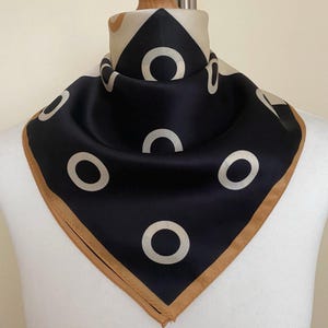 May include: A square scarf featuring a dark navy blue background with a repeating pattern of light beige circles. The scarf has a solid light beige border along the edges and is draped around a neck form to display the geometric print clearly.