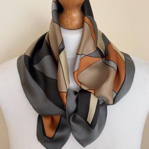 Puede incluir: Un pañuelo cuadrado con un patrón geométrico de líneas que se cruzan en tonos gris carbón, beige claro y naranja quemado. El pañuelo tiene un acabado suave y satinado, y está drapeado alrededor de un maniquí, mostrando un diseño abstracto.