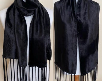 May include: A black scarf with a silky texture and long fringe. The scarf is draped over a mannequin, showcasing its length and the way it falls. The fabric has a subtle sheen, adding to its elegance.